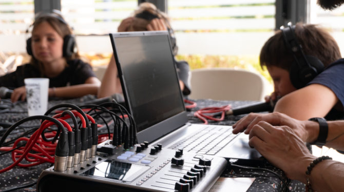 Photo d'une table de mixage au premier plan. Au second plan, un ordinateur et au dernier plan dans enfants qui parlent dans un micro